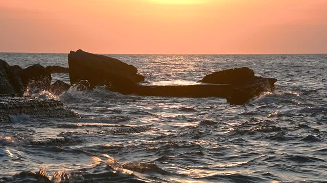 Dramatic sunset colors at sea. Waves hit the rocky shore and flysch cliff in Moon Bay, Slovenia. Reflection of yellow light on the surface. Real time, static shot, 4K