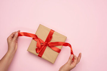 female hands untie a red ribbon on a gift wrapped in brown kraft paper on a pink background