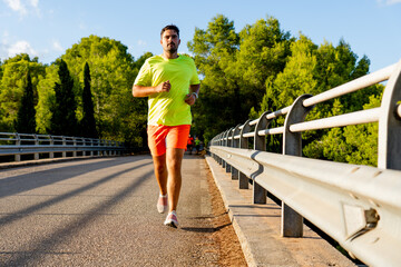 Young Spanish man with black hair and dark skin running on a bridge through a pine forest near the railing, wearing striking sportswear, yellow t-shirt and orange shorts