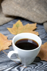 A cup of coffee among dried maple leaves. On pine planks painted black and white. Nearby is a rough linen cloth.