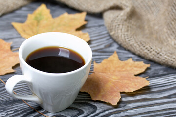 A cup of coffee among dried maple leaves. On pine planks painted black and white. Nearby is a rough linen cloth.
