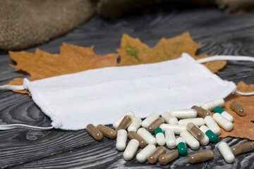 A handful of pills of different colors on a wooden one. Nearby is a medical mask. Among the dried up maple leaves. On a pine board surface.