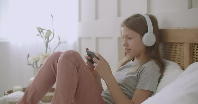 preteen girl is using app for communicating on smartphone, talking and listening by headphones, resting in her bedroom