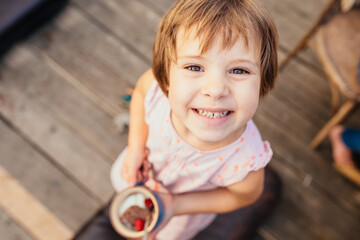 Top view of cute funny little girl with brown hair holding cup with dessert or ice cram. Portrait of cheerful little girl looking up in summer pink dress.