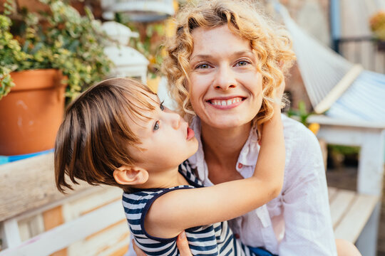 Blond curly excited happy woman and her son hugging together at garden or backyard. Cute little boy kissing her mother on terrace at summer day.