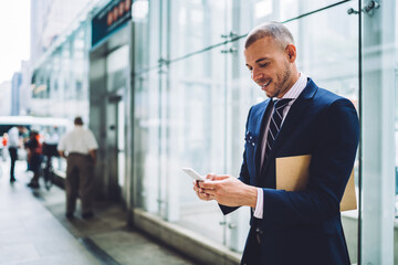 Joyful businessman with documents messaging smartphone in business hall