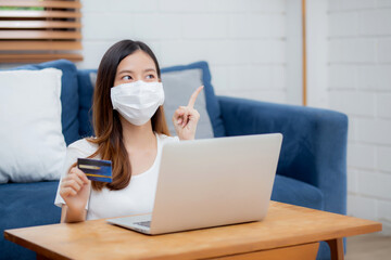 Young asian woman in face mask holding credit card shopping online with laptop computer buy and payment, girl using debit card purchase and thinking planning finance, lifestyle and new normal concept.