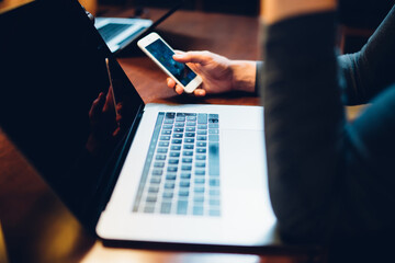 Person touch screening smartphone at laptop sitting in library