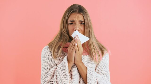 Female in cardigan and scarf is sneezing into handkerchief, she caught cold and looking ill while posing on pink background