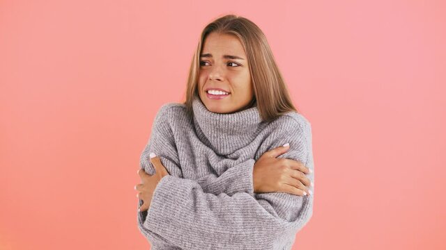 Lady in sweater is freezing, trying to keep warm rubbing herself by hands while posing against pink studio background