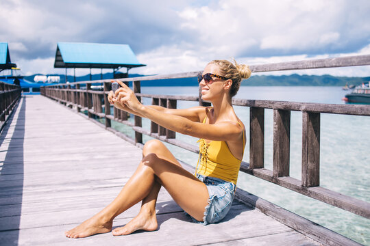 Happy hipster girl resting at Hawaii pier using cellular camera for clicking positive selfie pictures, carefree woman in sunglasses posing for smartphone while making media content on Raja Ampat