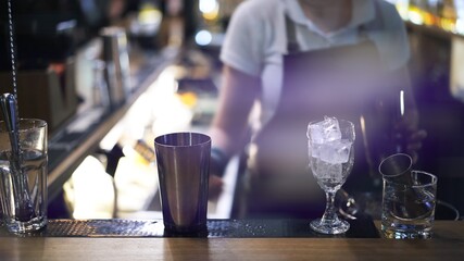 Metal cups and glasses on bar counter on background of bartender, lens flare. Bar counter with bar equipment for cocktail preparation, blurred background