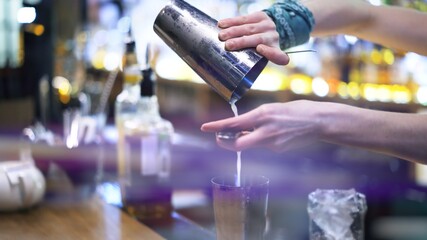 Bartender pouring alcohol through a strainer, lens flare. Cocktail pouring from stainless steel cup, cocktail making process, blurry background