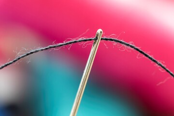 A close up portrait of some sewing thread put through the hole of a needle. The yarn is black and the background is out of focus and colorful. The needle is ready for some needlework or sewing.