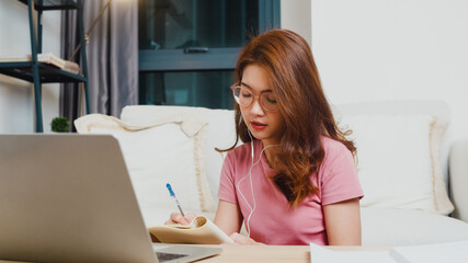 Young Asia teen girl student wears headphones distance learning lesson with online teacher on laptop in living room from home at night. Social distancing, quarantine for corona virus prevention.