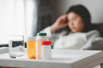 Medicines and glass of water with Asian woman feeling sick and headache in background in living room