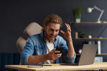Angry young handsome curly man using laptop. Stressed freelancer lost data on broken not working laptop. Receiving bad news or email, spam message. Young people working with mobile devices.