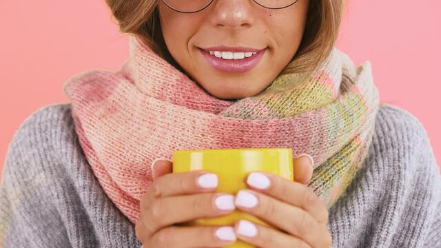 Unknown woman in warm sweater, scarf and glasses is holding cup of tea or coffee inhaling its aroma, smiling posing on pink background