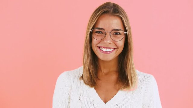 Young lady in glasses and white jumper is pointing at you by forefingers, winking and laughing posing on pink background
