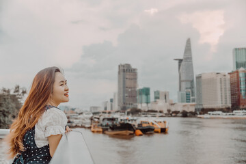 Fototapeta na wymiar a Vietnamese girl standing by the riverbank admiring the view of Ho Chi Minh city with Bitexco Financial Tower, many buildings and Saigon river in blurred background.