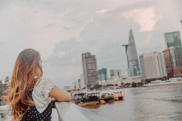 Plakat a Vietnamese girl standing by the riverbank admiring the view of Ho Chi Minh city with Bitexco Financial Tower, many buildings and Saigon river in blurred background.