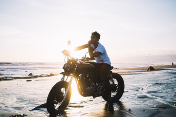 Obraz na płótnie Canvas Hispanic man sitting on motorcycle on sea beach and pondering during sunset