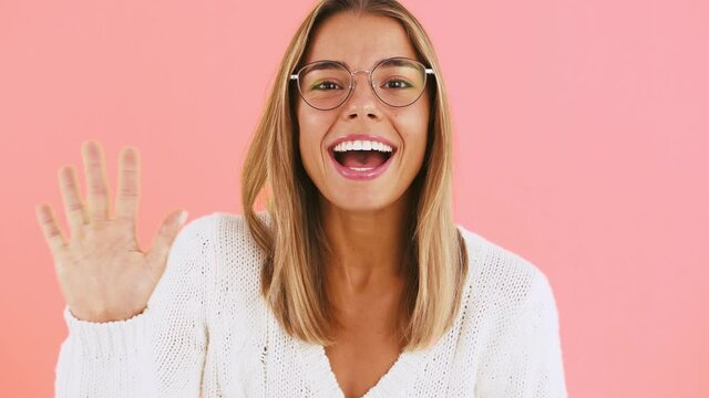 Young female in glasses and white jumper is waving her hands, saying hi and smiling while posing on pink background