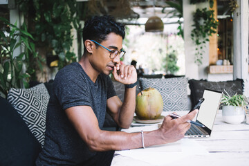 Thoughtful young ethnic student using cellphone at table at cafe