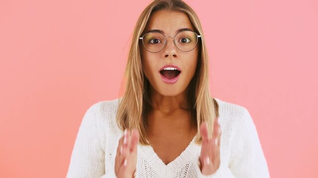 Young lady in glasses and white jumper is looking surprised, saying wow and smiling while posing on pink background