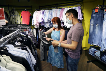 Young married couple wearing medical face masks are choosing clothes in the store. A new reality with health care