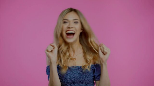 Excited blonde woman dancing and showing yes gesture isolated on purple