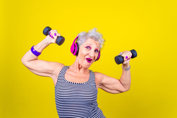 Elderly woman training using dumbbells on yellow background - Isolated mature woman stretching using gym weight listening music