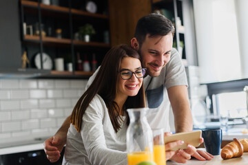 Smiling couple enjoying a beautiful morning at home kitchen, having breakfast, drinking coffee and juice, and looking at smartphone