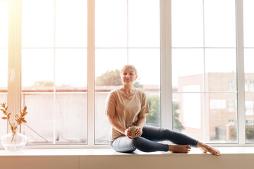 Fototapeta premium Beautiful daydreamer. Beautiful young woman holding coffee cup, smiling while sitting at windowsill at home