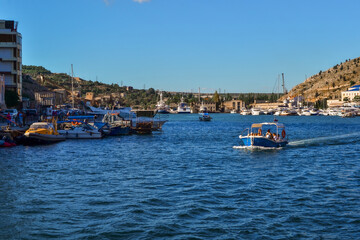 Obraz premium bright blue boat along the bay of resort town of Balaklava among white ships on docks near houses and buildings, sunny