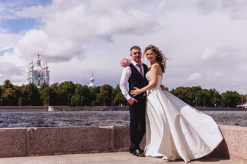 Bride and groom stand embracing on background of river and Cathedral. Newlyweds in wedding dresses on Sunny wedding day. Couple on nature in amazing view. Newlyweds in love happy together