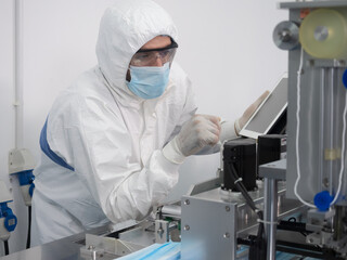 Naklejka premium The engineer worker in protective suits, masks, and gloves is checking and controlling the machine in the manufacture of surgical masks for coronavirus..