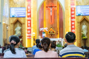 A blurred background photo of the inside of a Vietnamese church sanctuary that is filled with people in the pews, and the pastor stands under a large cross at the altar, in Vietnam.