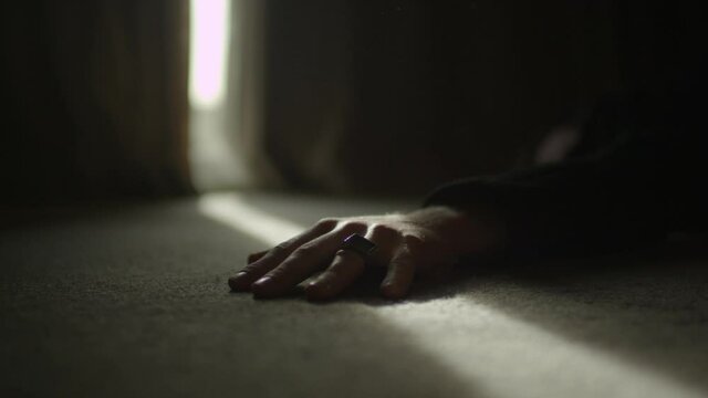 A person's hand lies motionless on the carpeted floor as a shard of light shines in through the curtains