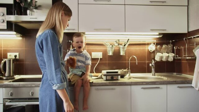 Young mother and her son are making waffles in waffle maker at home kitchen. Little boy sits on the table and eats the waffle