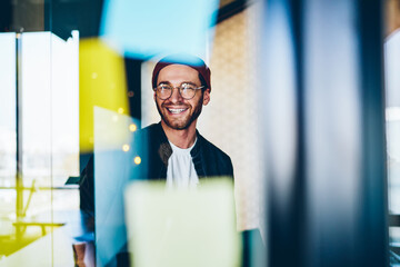 Fototapeta Cheerful caucasian hipster guy in trendy eyewear satisfied with creative job laughing sitting near stickers with ideas in office, 20s smiling carefree male student enjoying learning in business school obraz