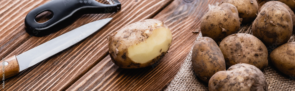 Wall mural dirty potatoes on wooden table with peeler and knife, panoramic shot