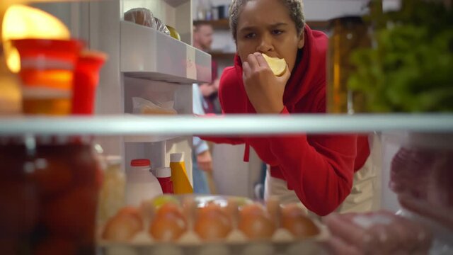 African Woman Eating Apple And Taking Yoghurt From Fridge At Home Kitchen