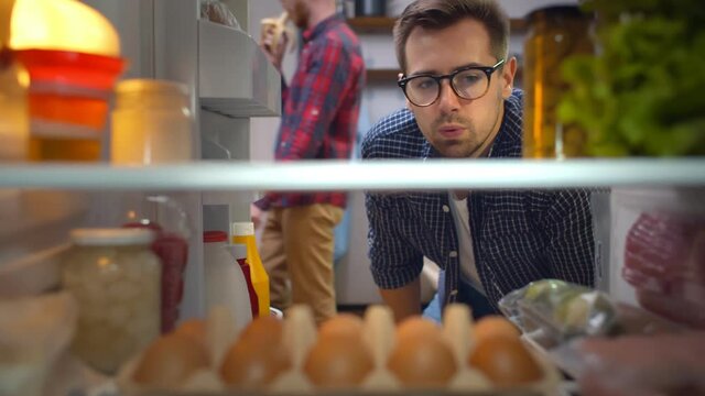 Young guy opening fridge looking for snack checking time on wristwatch and grabbing healthy yogurt