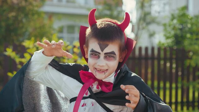 Portrait shot of cute little boy wearing black and white makeup and devil horns hiding his face behind cape, then making scary face for camera outdoors