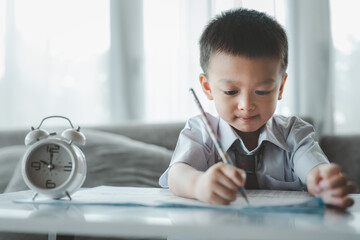 Naklejka premium Asian Boy Doing homework with the intention. Child boy holding pencil writing, A boy drawing on white paper at the table,Elementary school and home schooling, Distance Education concept.