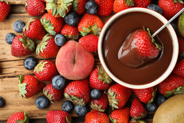 Fondue fork with strawberry in bowl of melted chocolate surrounded by other fruits on wooden table, flat lay