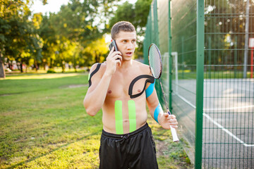 Young caucasian professional tennis player with kinesiology taping on body carrying racket near fenced sports ground. Confident man with muscular naked torso talking using smartphone.