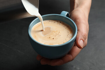 Woman pouring milk into cup of coffee at black table, closeup