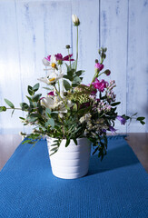 multicolored wildflowers stand on a wooden table, on a blue cloth napkin, in a white vase against a blue wall   